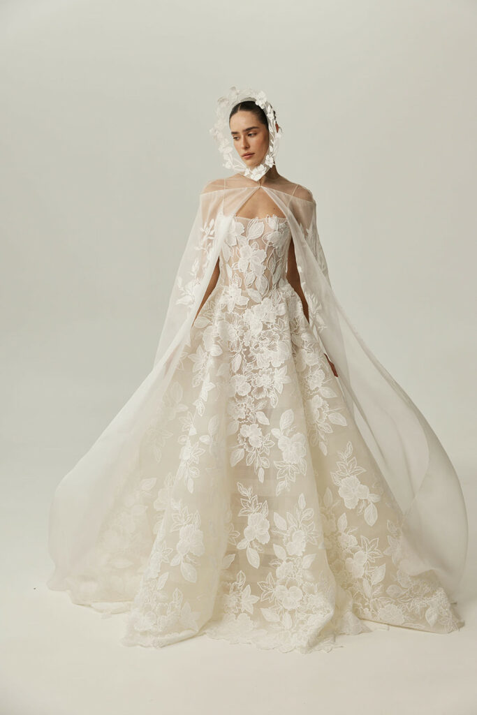 Woman in sheer lace bridal gown, cape, headpiece.