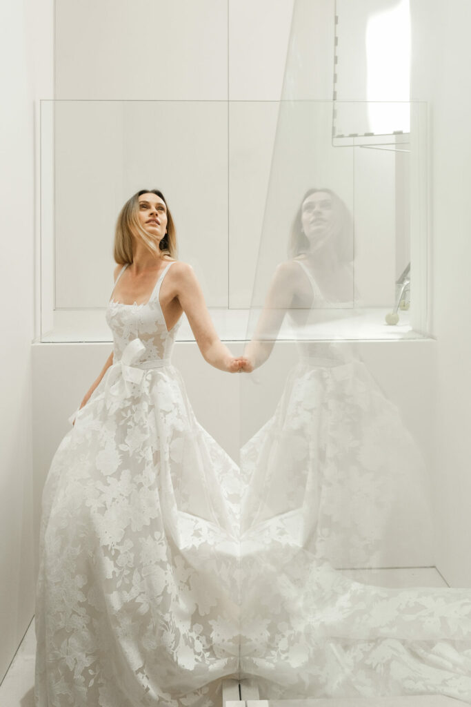 Woman in lace wedding gown stands by glass, reflecting.