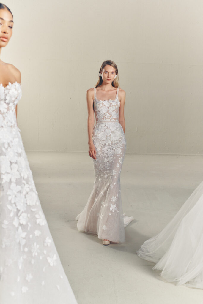 Model in beige sheer gown, floral details, plain backdrop.