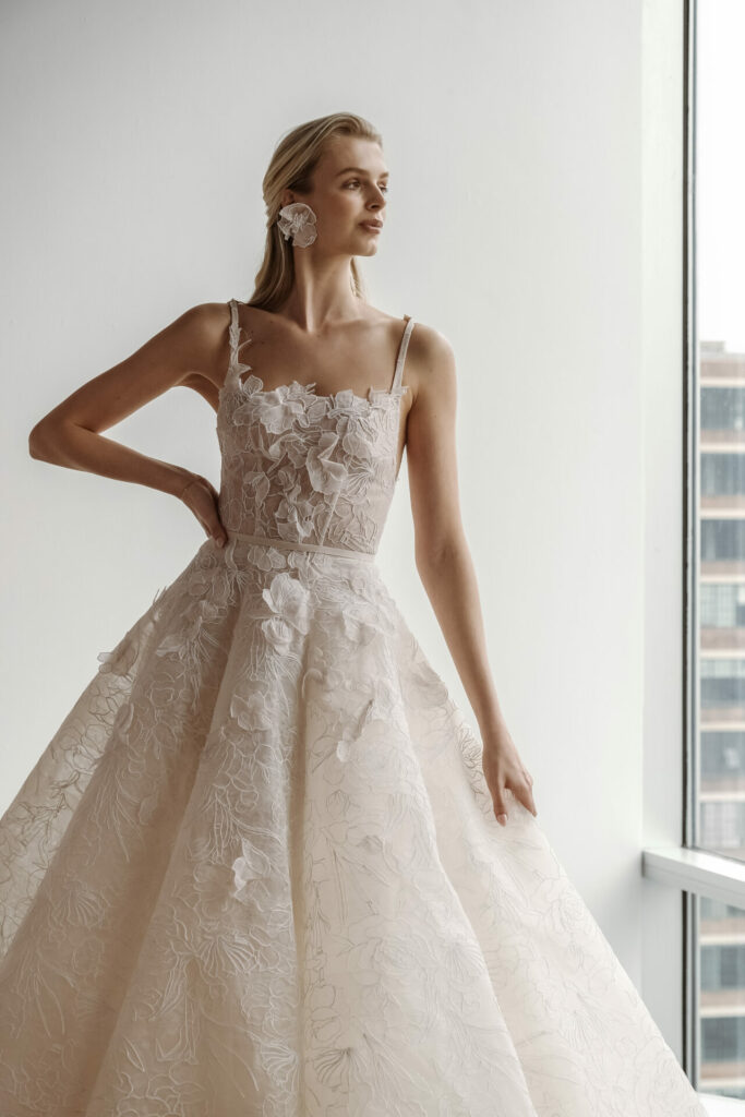 Woman in blush pink floral gown poses by window.