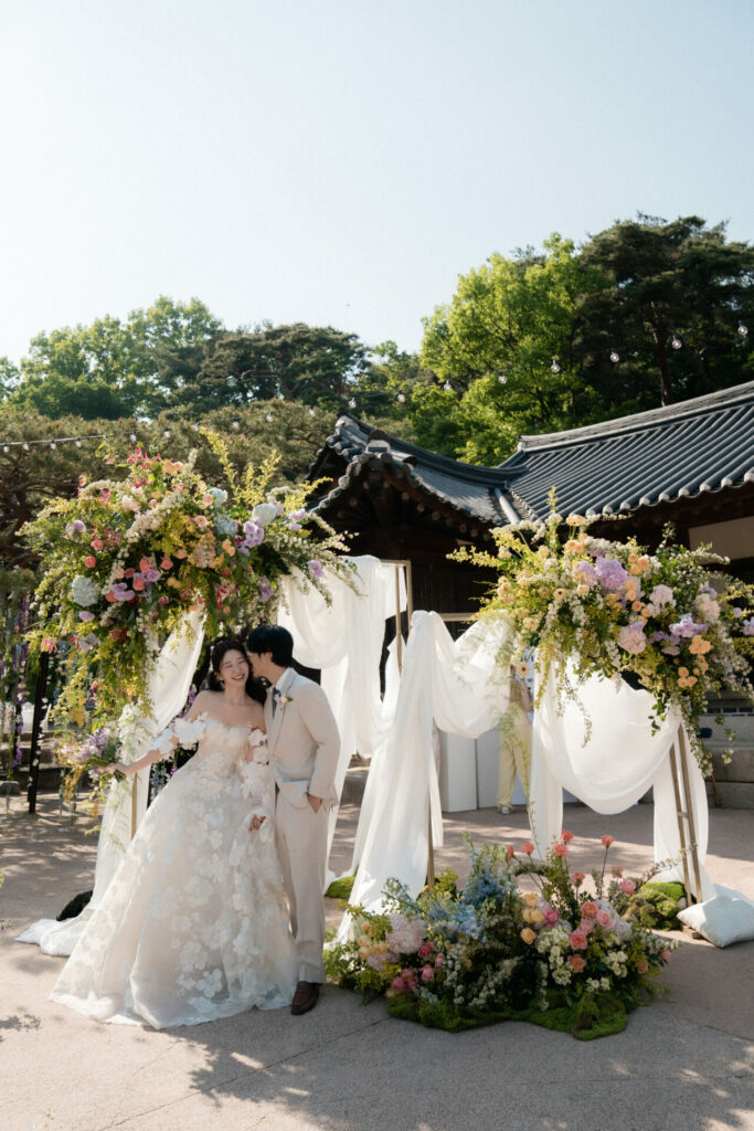 Groom kisses bride’s cheek under floral arches outdoors.