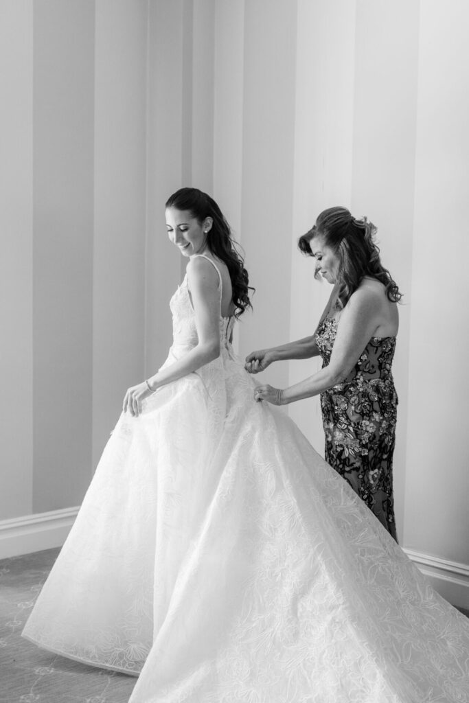 Woman fastens smiling bride’s couture gown in soft light.