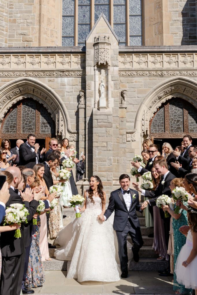 Bride and groom exit church in sunlight, cheered.
