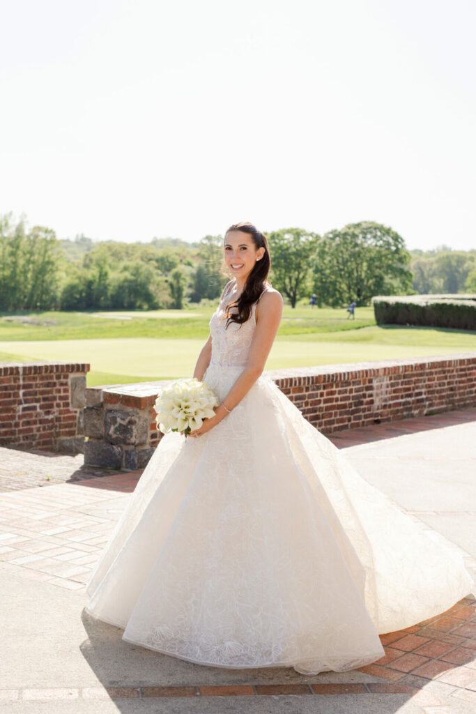 Katya Ruiz in white gown outdoors, greenery and brick backdrop.