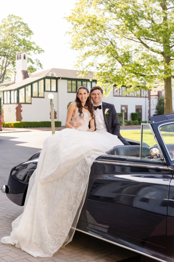 Bride and groom smiling by classic car, elegant backdrop.