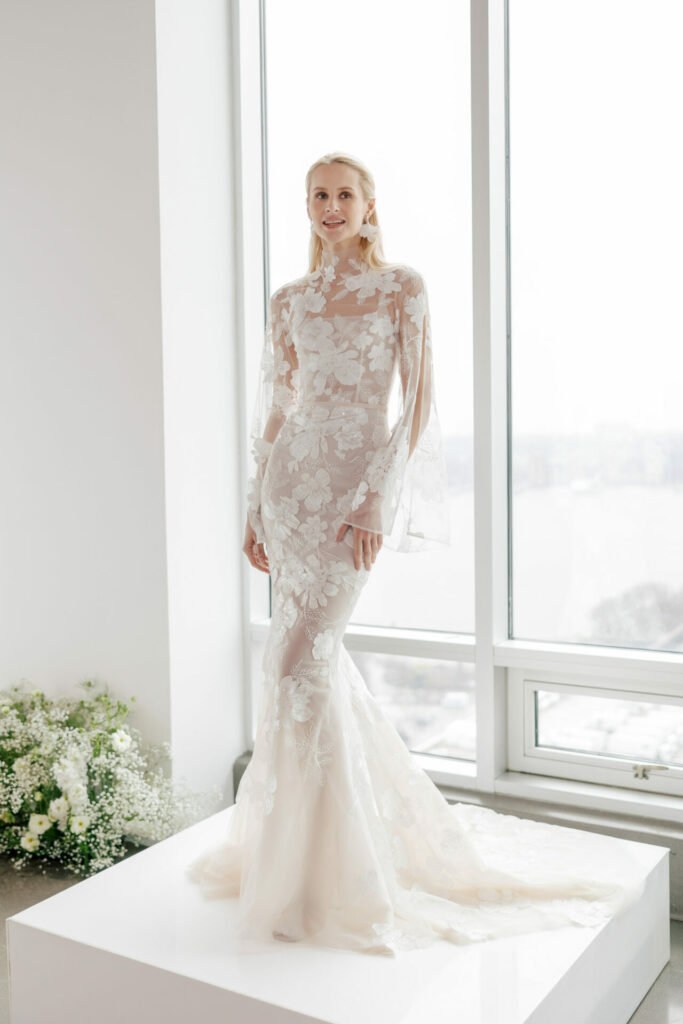 Woman in white lace dress by large bright windows.