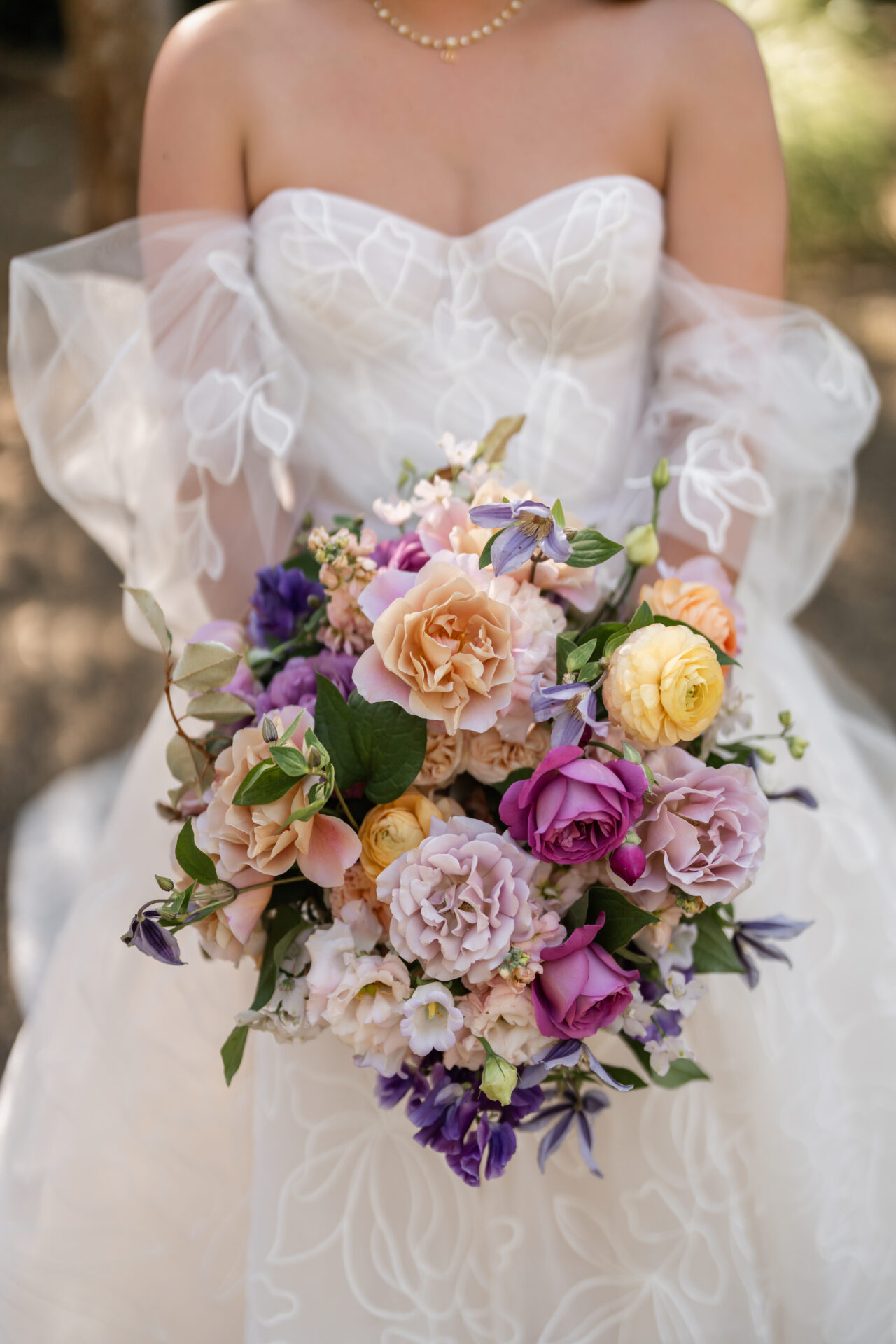 Bride in off-shoulder white gown holding pastel bouquet outdoors.