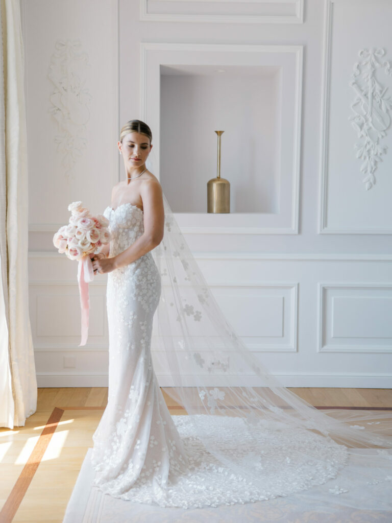 Bride in lace gown holds bouquet indoors, floral veil.