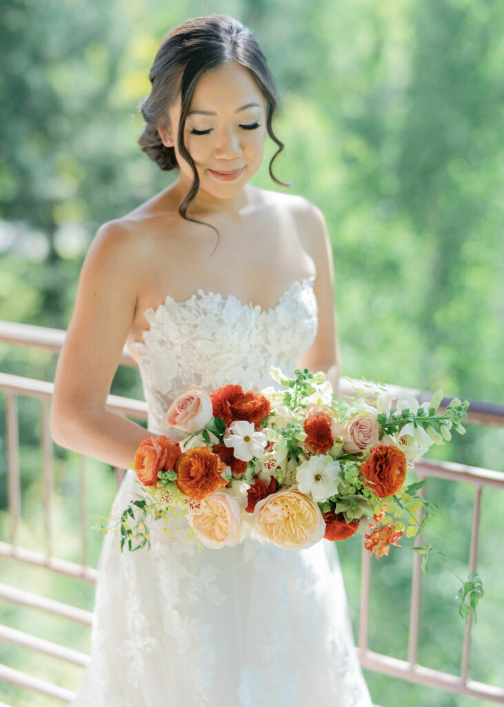 Bride in white lace gown holds colorful bouquet outdoors.