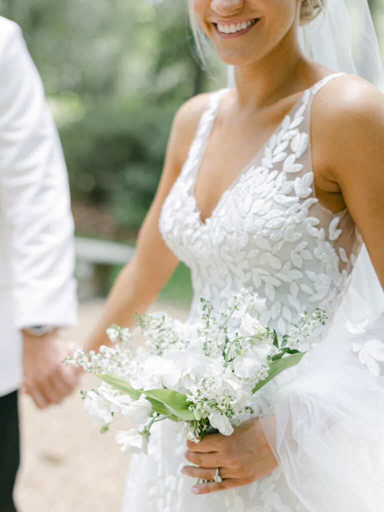 Bride in lace dress smiles, holding bouquet outdoors.
