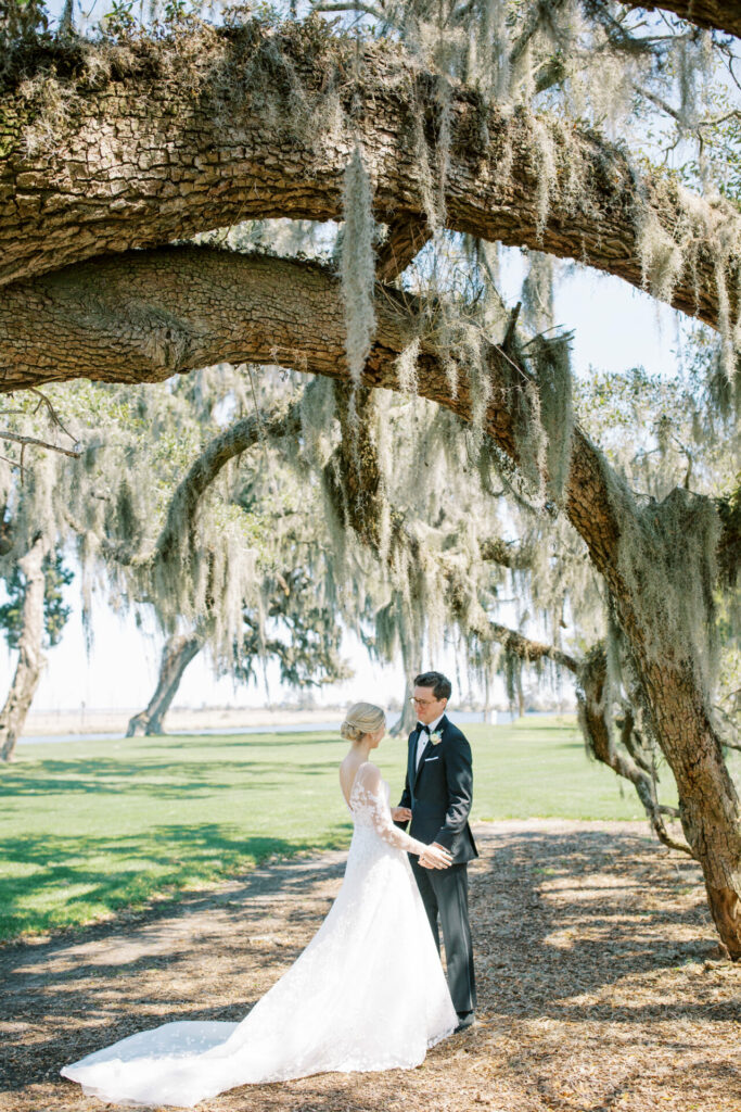 Bride and groom hold hands under sunlit, mossy oak.
