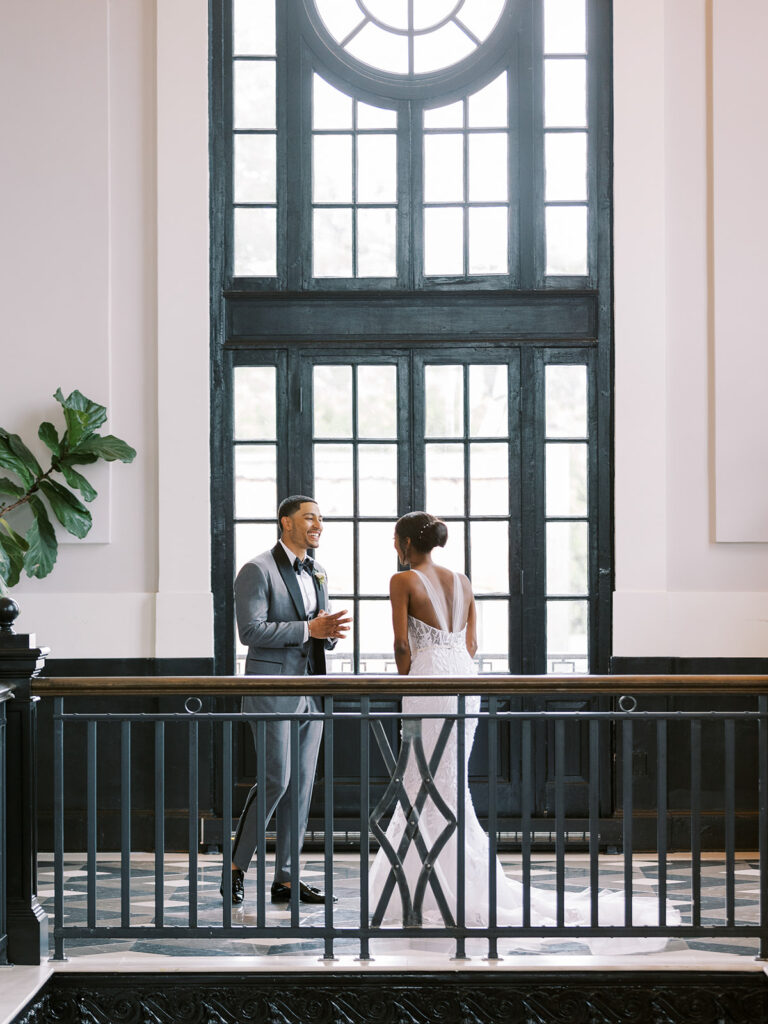 Bride and groom smile, facing each other indoors.