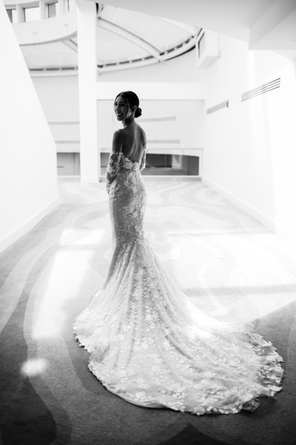 Bride in lace gown poses in sunlit hallway.