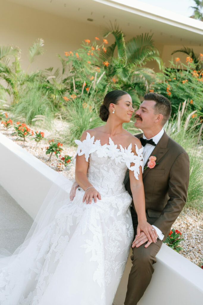Katya Ruiz in floral gown with groom, surrounded by lush greenery and orange flowers.
