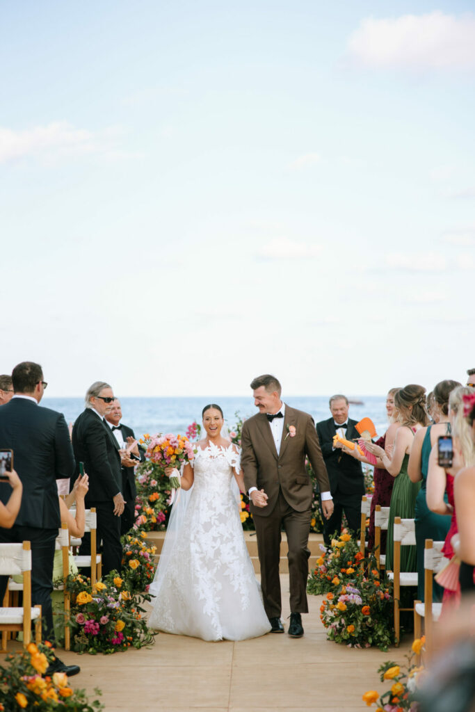 Bride and groom walk aisle by ocean, guests watching.