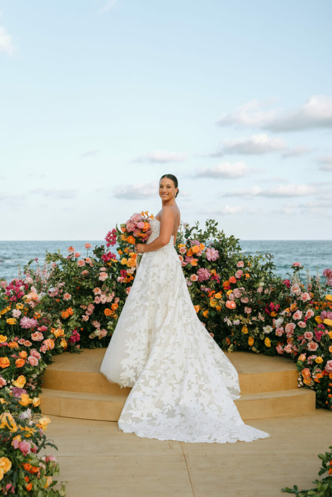 Bride in couture gown on steps amid flowers, ocean backdrop.