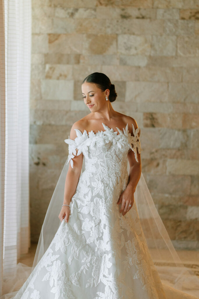 Bride in off-shoulder gown smiles, sleek bun, veil.