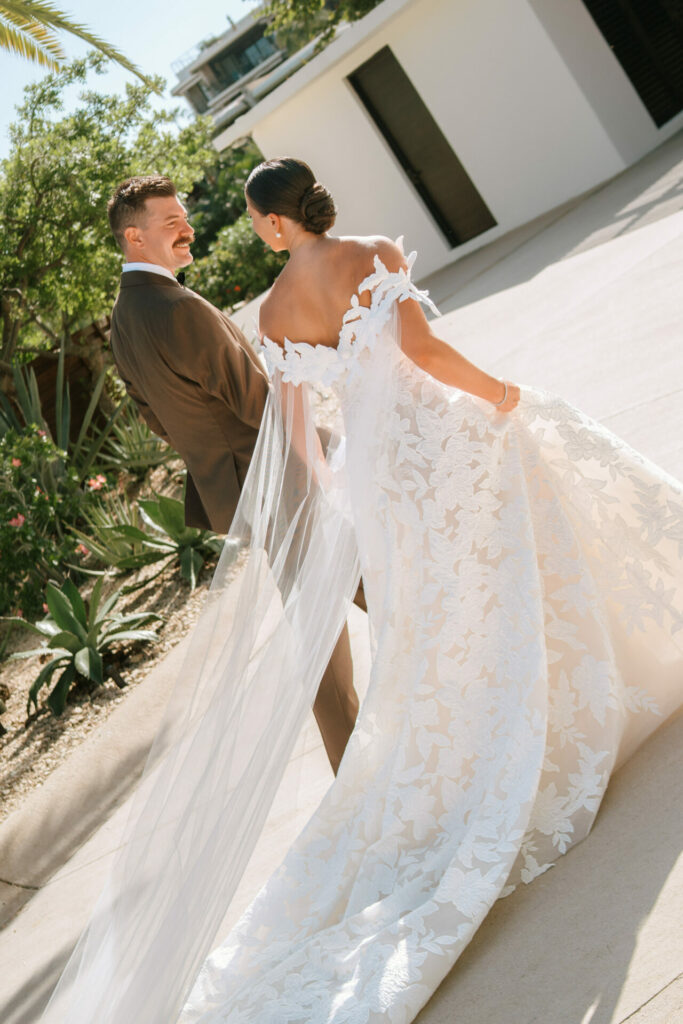 Bride and groom smiling, walking outdoors near greenery.