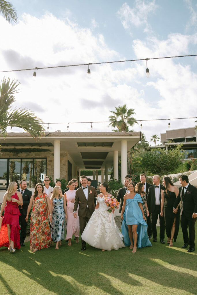 Katya Ruiz and groom walk hand-in-hand under glowing lights, friends nearby, her Mira Zwillinger gown shining among swaying palms.