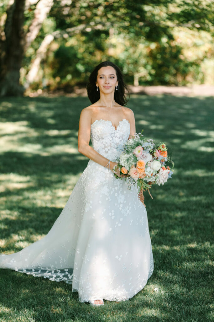 Bride in floral strapless gown holds bouquet outdoors in sunlight.