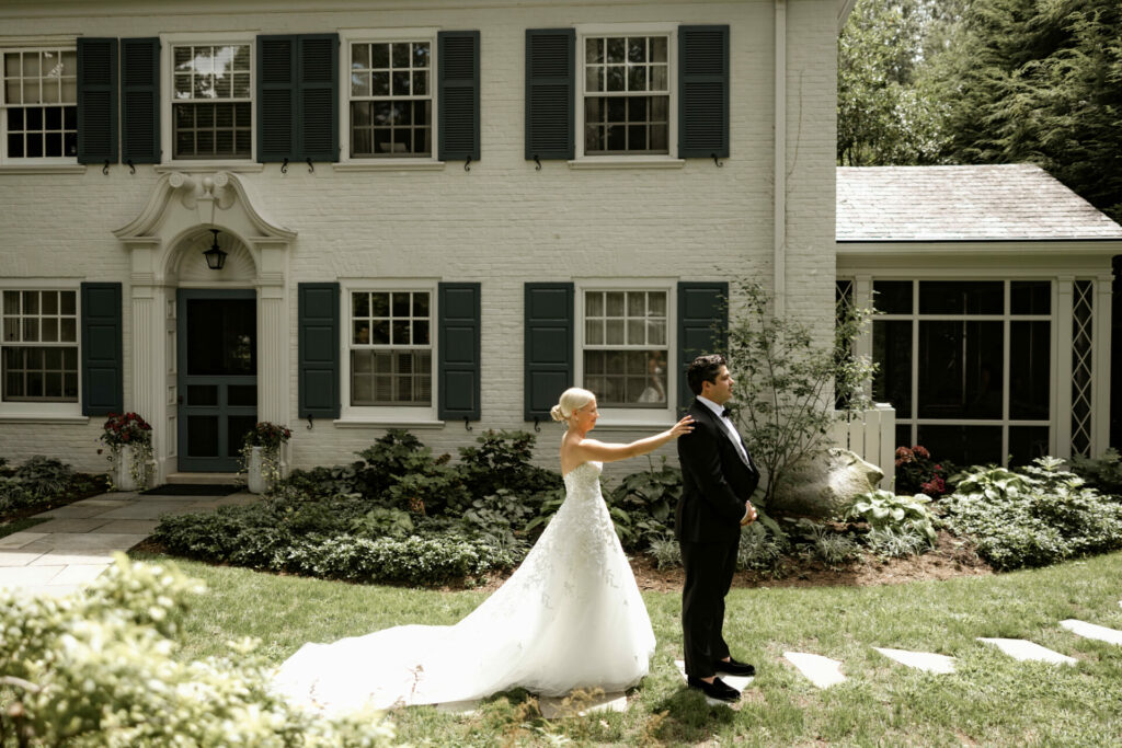 Bride in white gown behind groom outside blue-shuttered house.