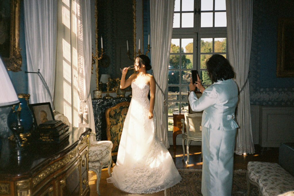 Bride in gown brushes teeth as friend photographs.