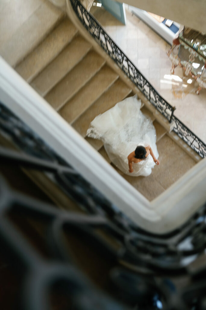 Bride in Mira Zwillinger gown ascends curved staircase.