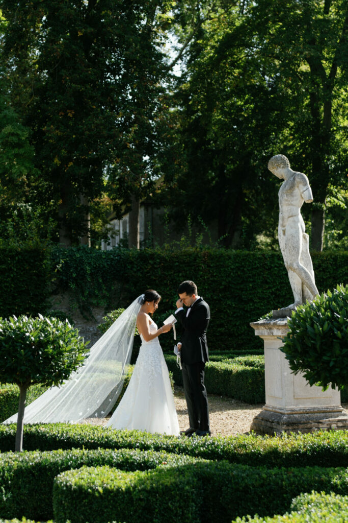 Katya Ruiz and groom in garden, elegant attire, statue nearby.