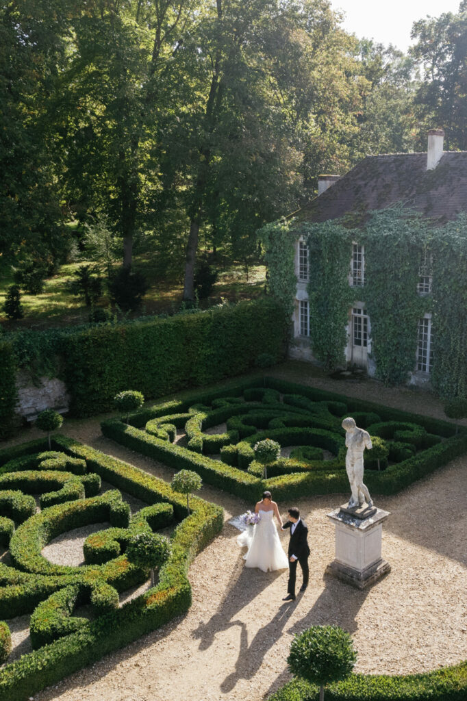 Bride in Mira Zwillinger gown walks garden with groom.