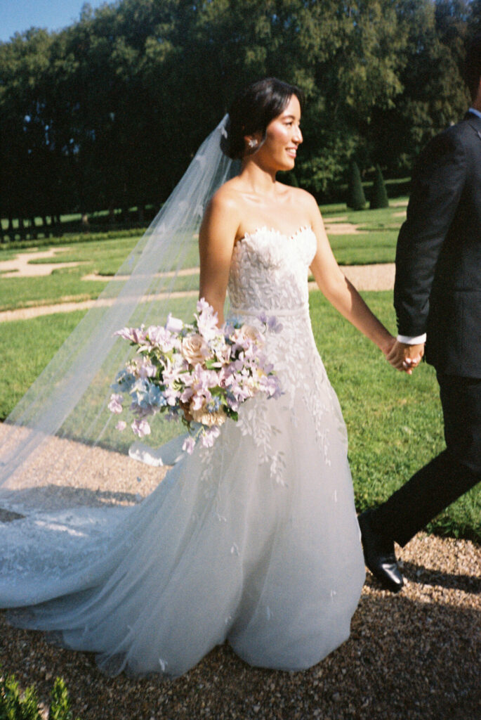 Katya Ruiz in elegant bridal gown walks with suited man outdoors.