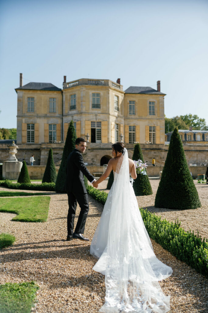 Bride and groom stroll garden by yellow chateau.