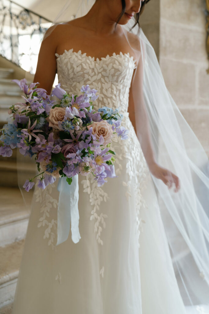 Katya Ruiz in leaf-patterned gown, lilac bouquet, veil trailing by stone stairs.