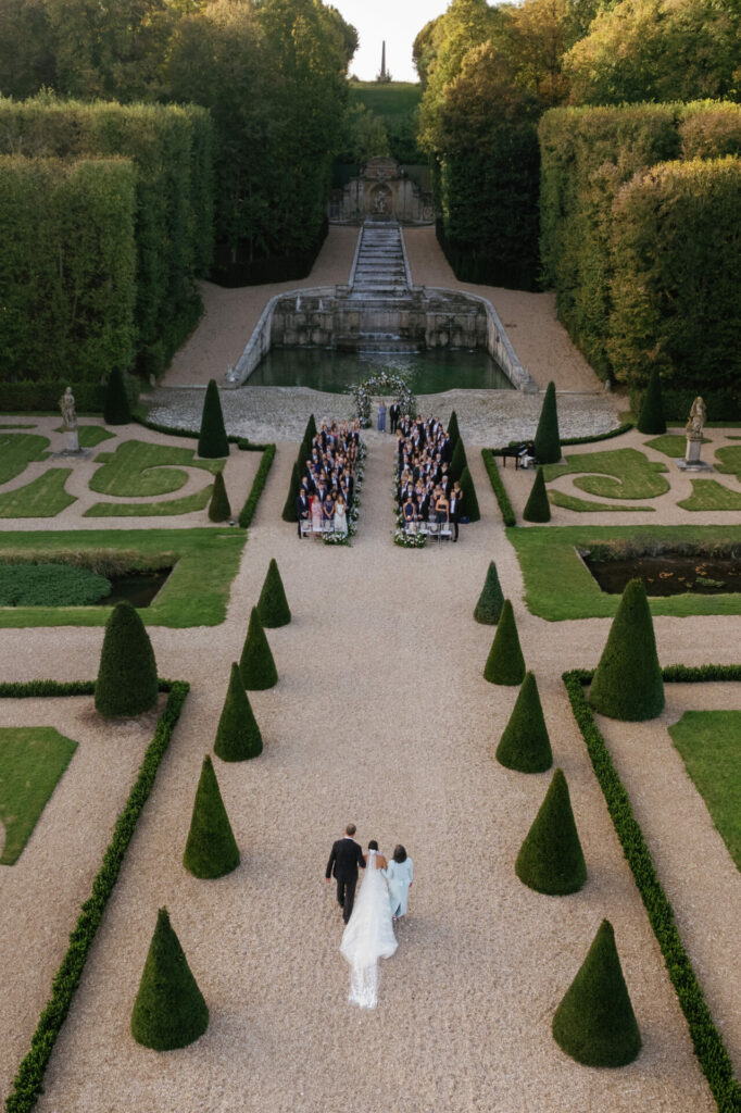Bride and groom walk garden path to outdoor ceremony.