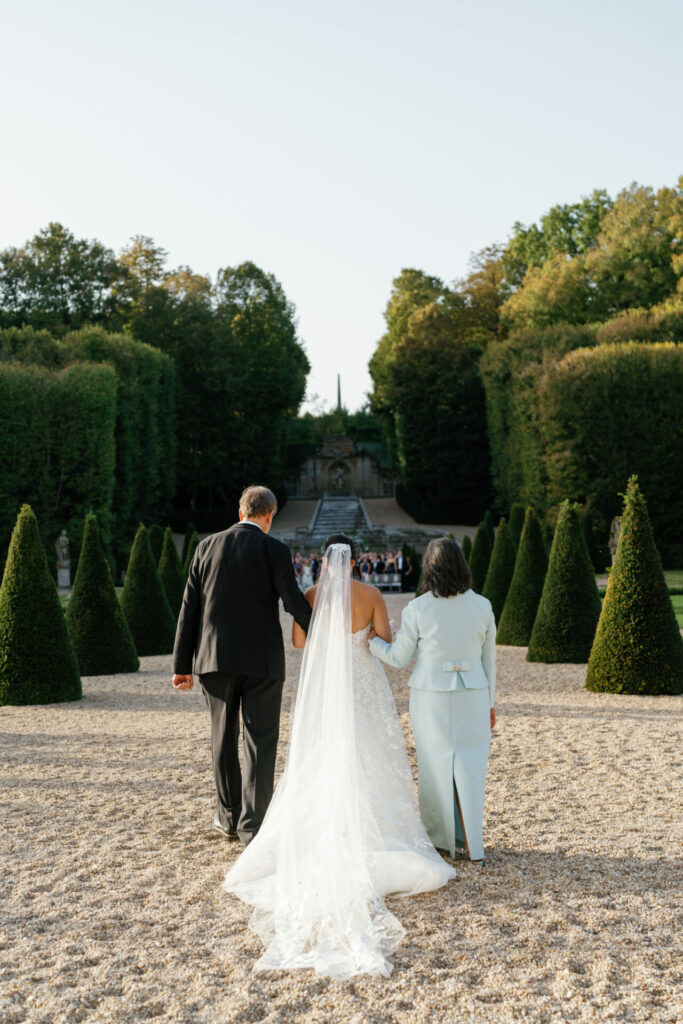 Bride in Zwillinger gown walks garden aisle with parents.