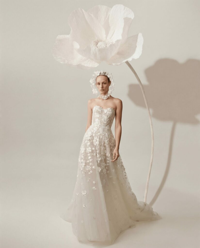 Serina in floral white gown beside giant flower, shadowed.