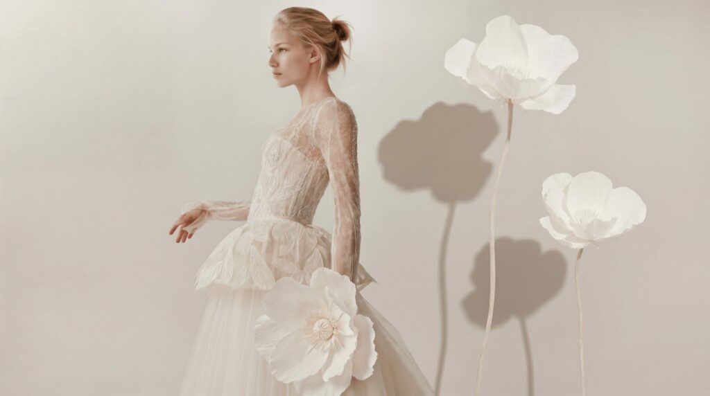 Ena in lace dress holds flower, soft shadows nearby.