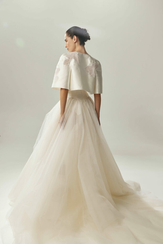 Woman in white tulle gown, floral cape, facing away.