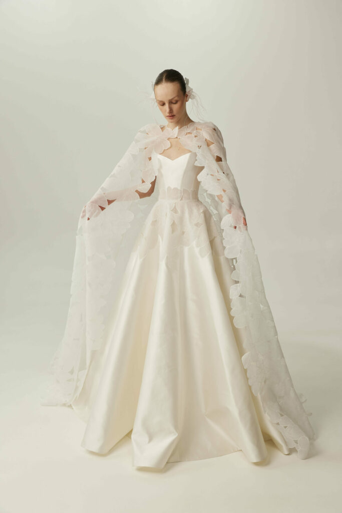 Serina in white gown, holding floral cape open.