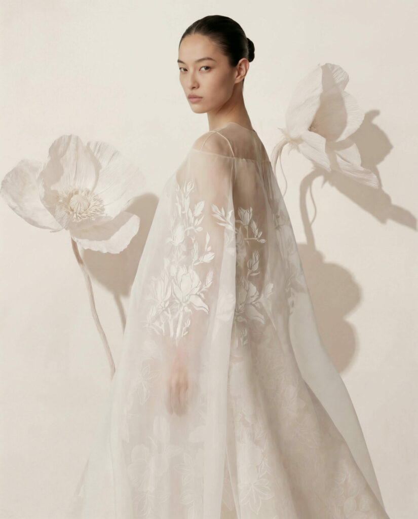 Woman in sheer white sui dress before giant paper flowers.
