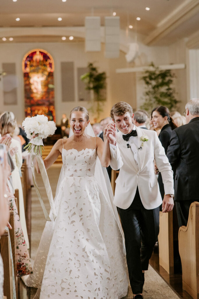 Smiling newlyweds walk aisle; guests admire bride’s couture.