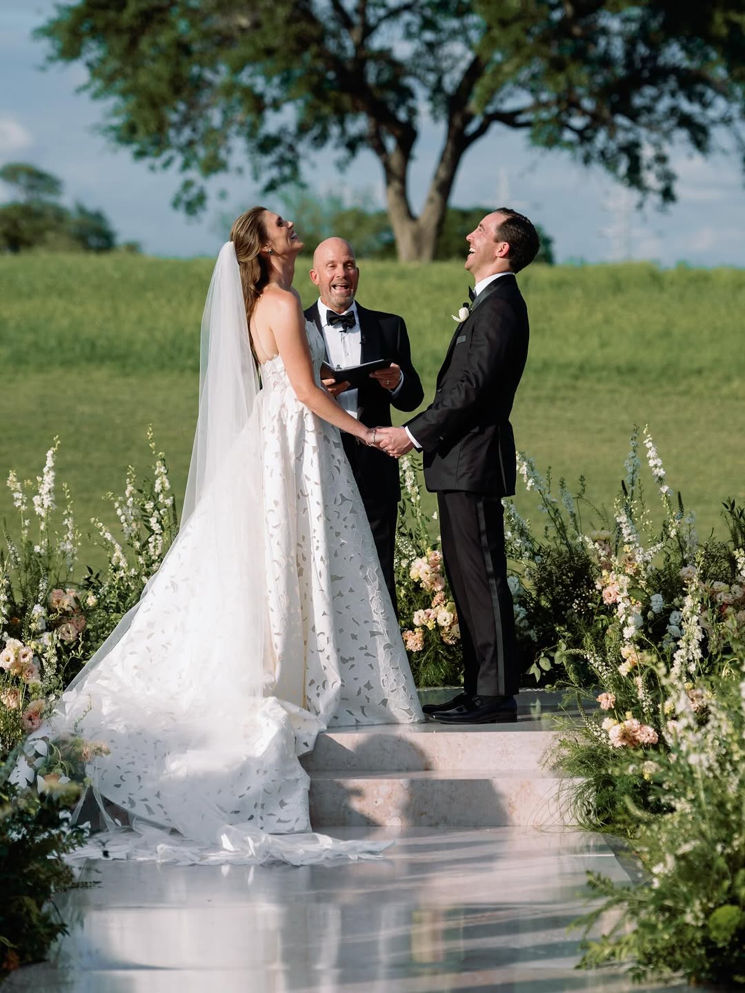 Bride and groom hold hands outdoors, officiant smiles.