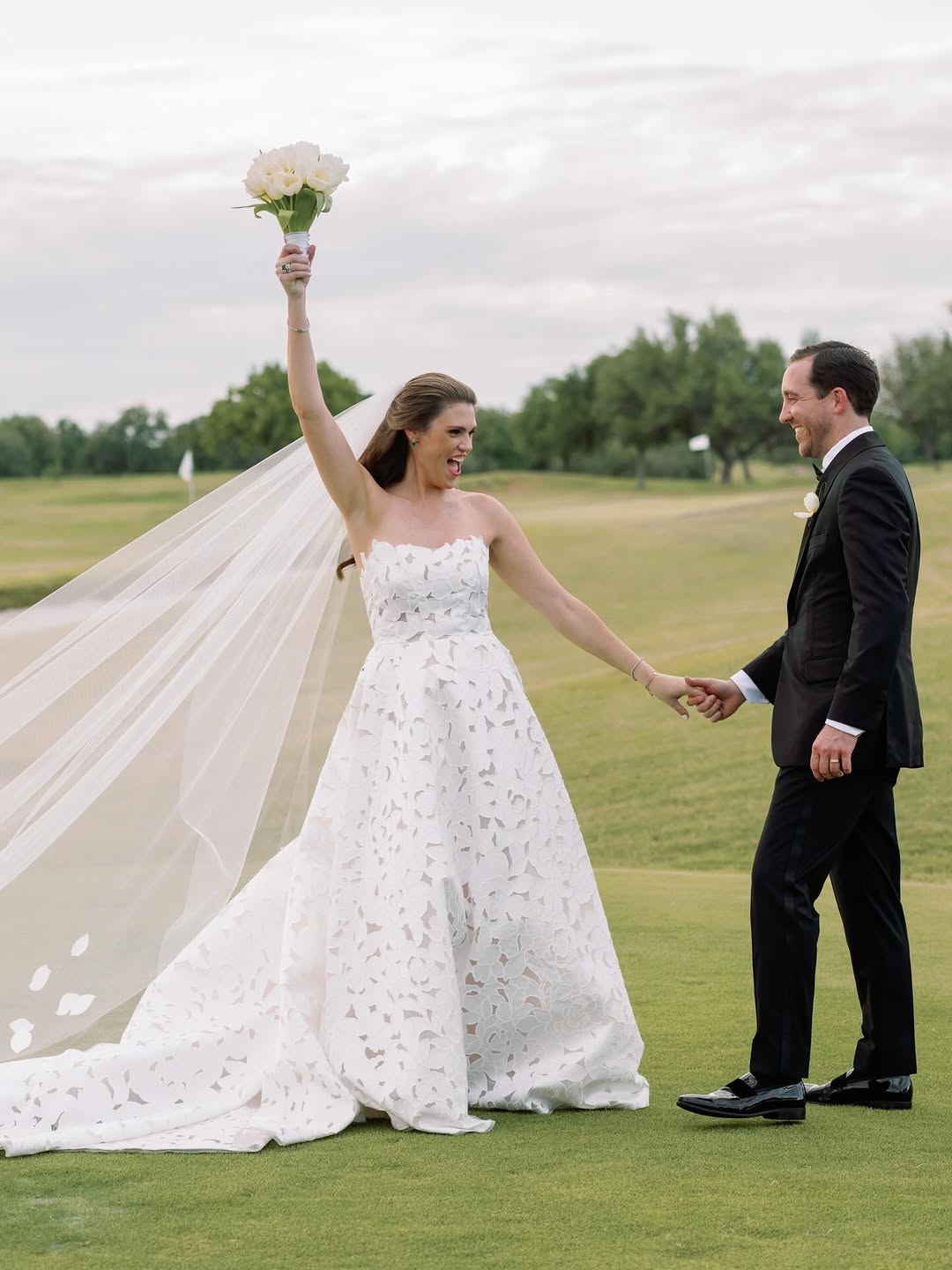 Bride triumphantly lifts bouquet beside smiling groom outdoors.