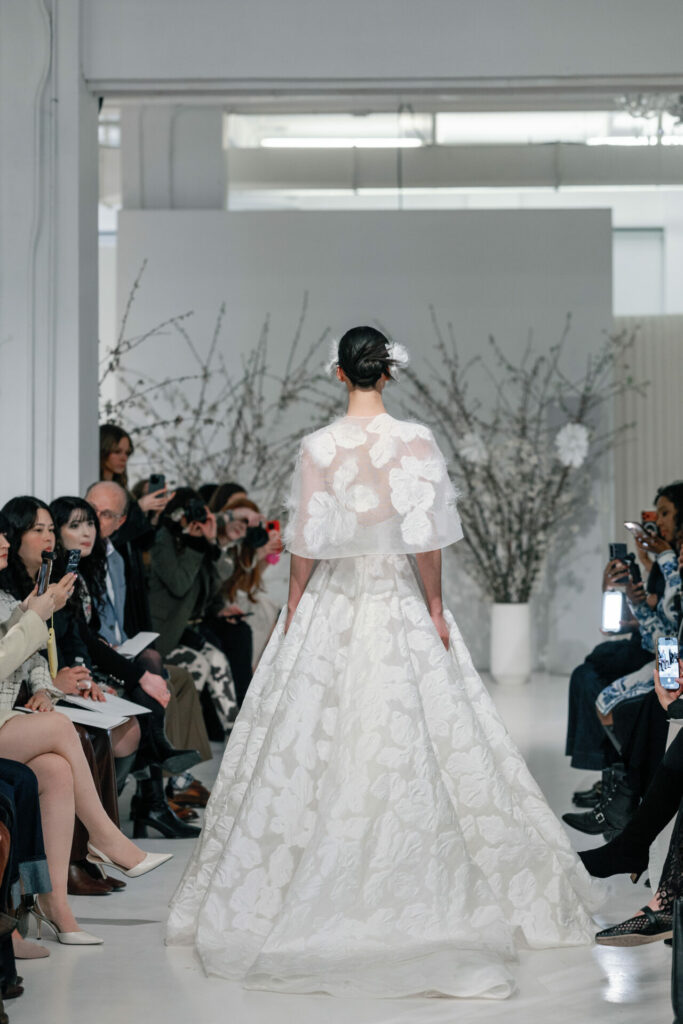 Model in white floral gown and cape on runway.