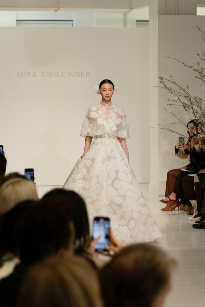 Model in floral Clea wedding dress walks runway; audience photographs; backdrop reads Mira Zwillinger.