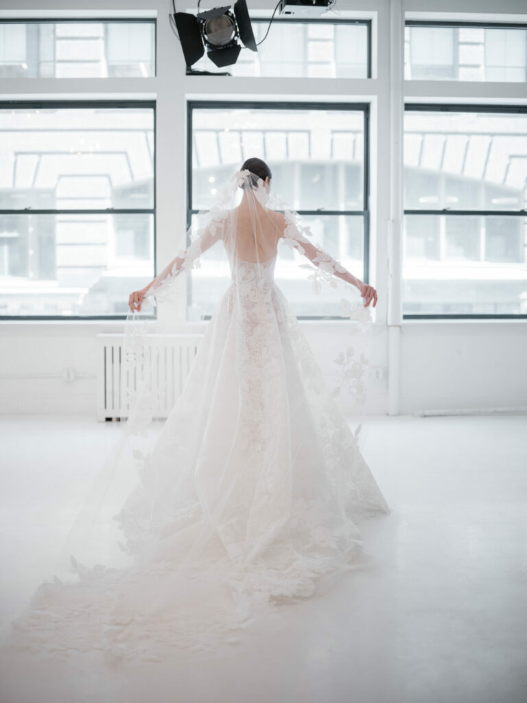 Bride in lace gown holds veil by bright windows.