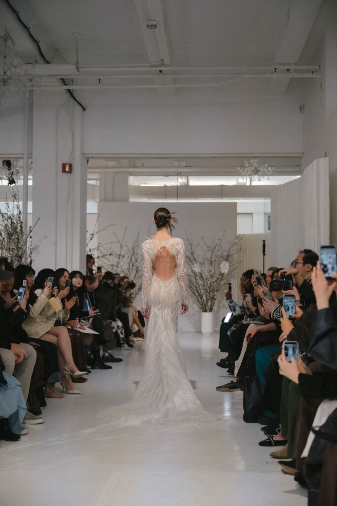Model in sheer white gown walks runway, audience photographing.