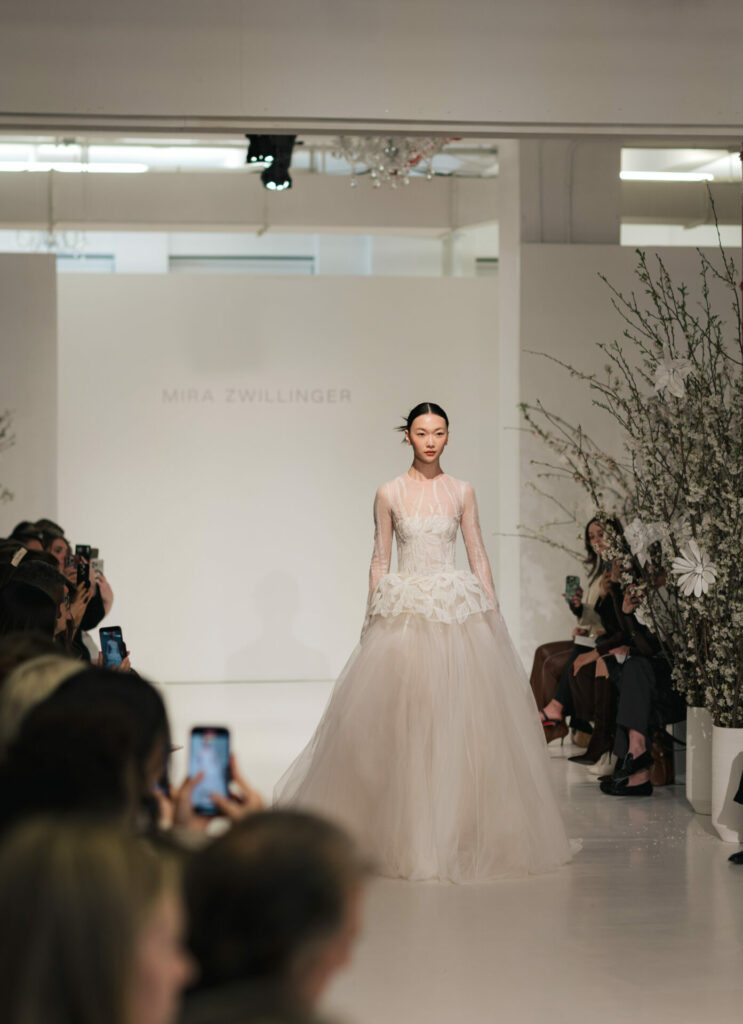 Model in white lace gown on runway; floral backdrop.