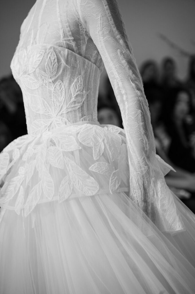 Model in lace wedding dress; blurred audience behind.