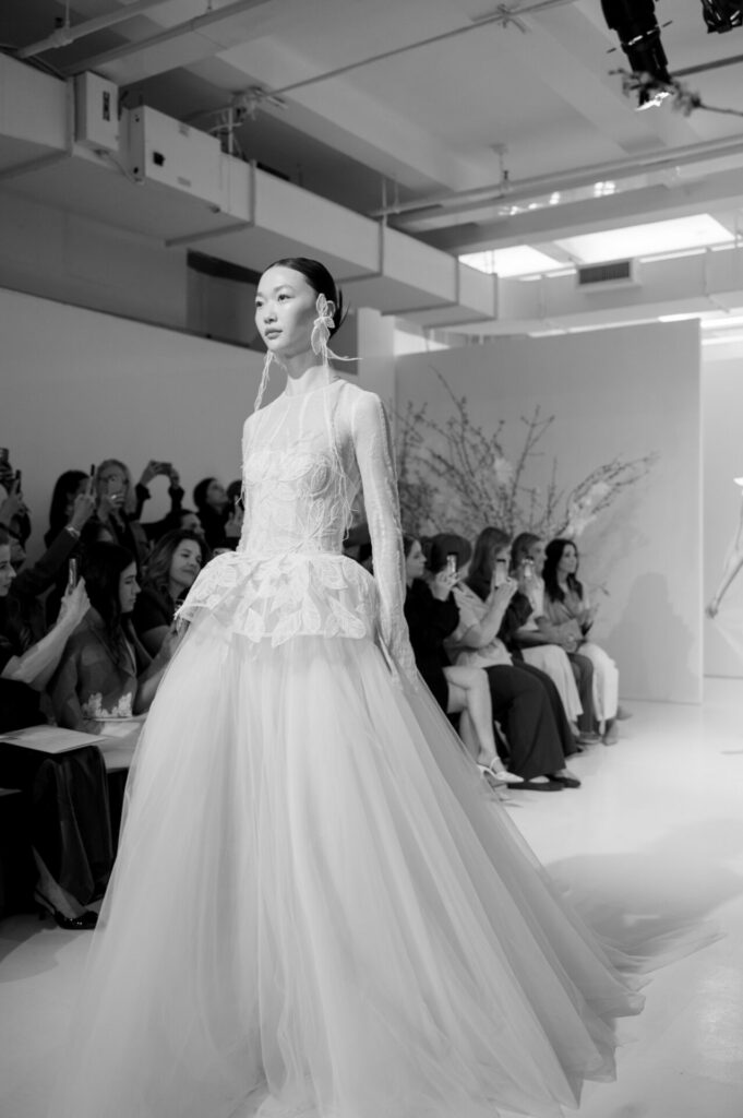 Model in lace tulle bridal gown walks runway.