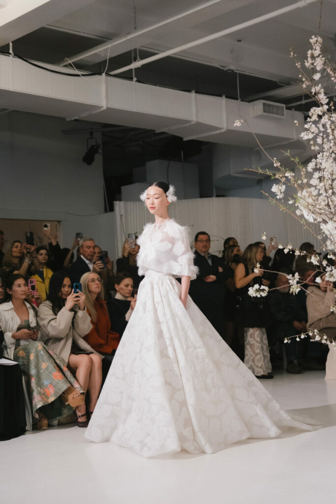 Model in white floral Clea dress walks runway; audience watches.